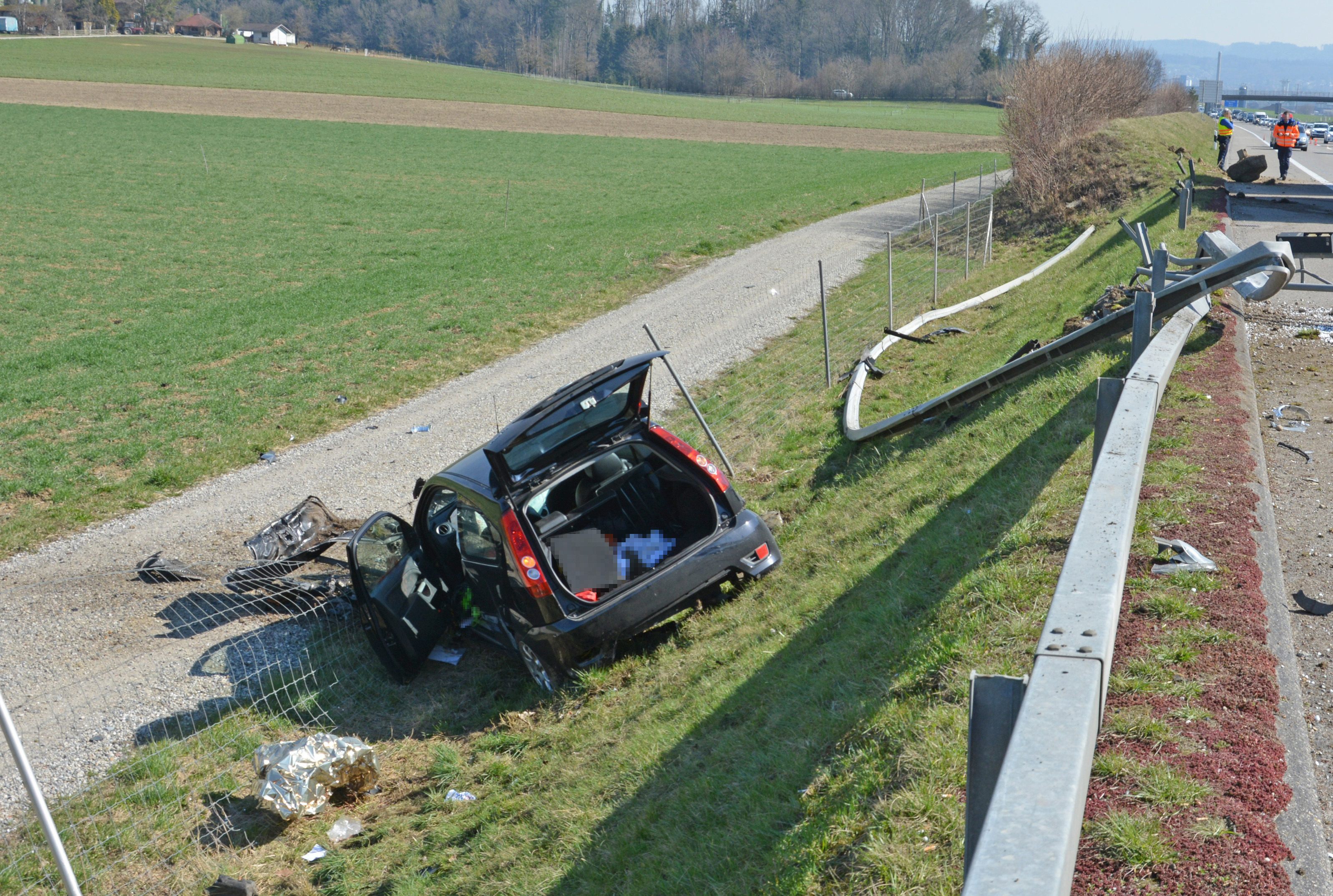 Stark beschädigte Autobahneinrichtung und Fahrzeug auf der Seite liegend im Wildzaun.